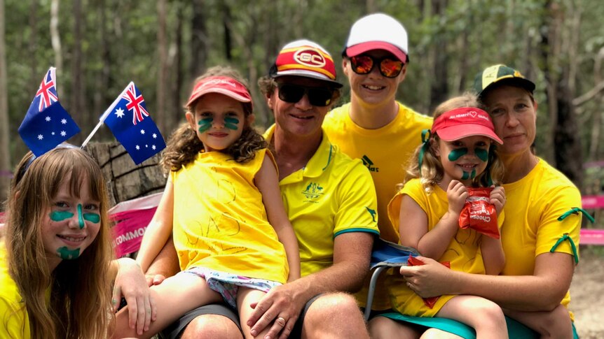 The six members of the Williams family sit together in their green and gold with the mountain biking track behind them