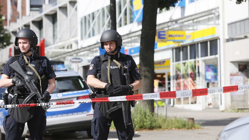 Heavily armed police officers secure the area after a knife attack in Hamburg, standing in front of red and white police tape.