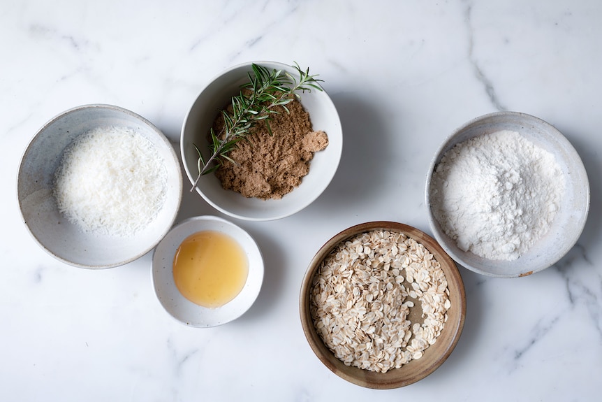 Anzac biscuit ingredients laid out in bowls with desiccated coconut, brown sugar, rosemary, honey and plain flour, ready to bake