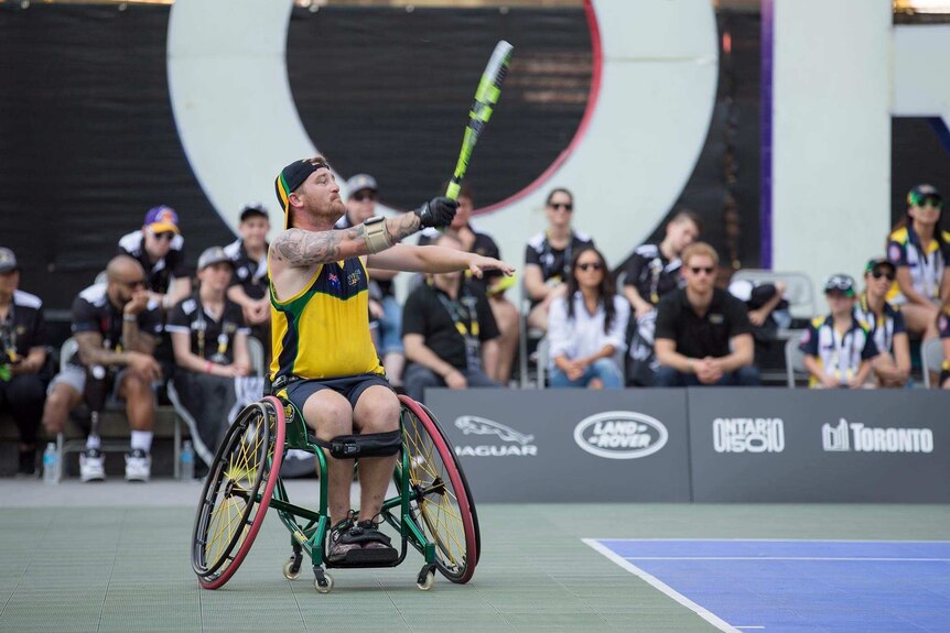 Wheelchair tennis player serves a ball as crowd looks on in the background.