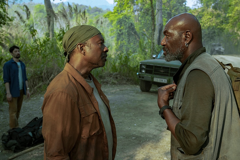 Two men in casual clothes talk face to face on road near static car and green jungle, behind them a man in blue shirt watches.