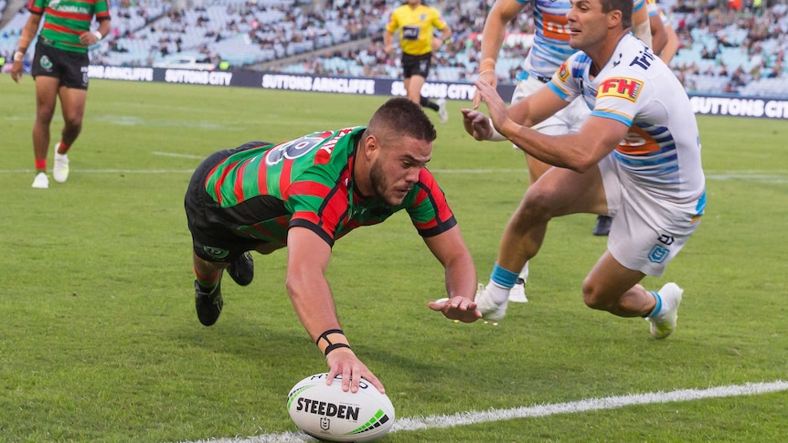 An NRL player dives to plant the ball in the corner for a try, as defenders try to grab him.