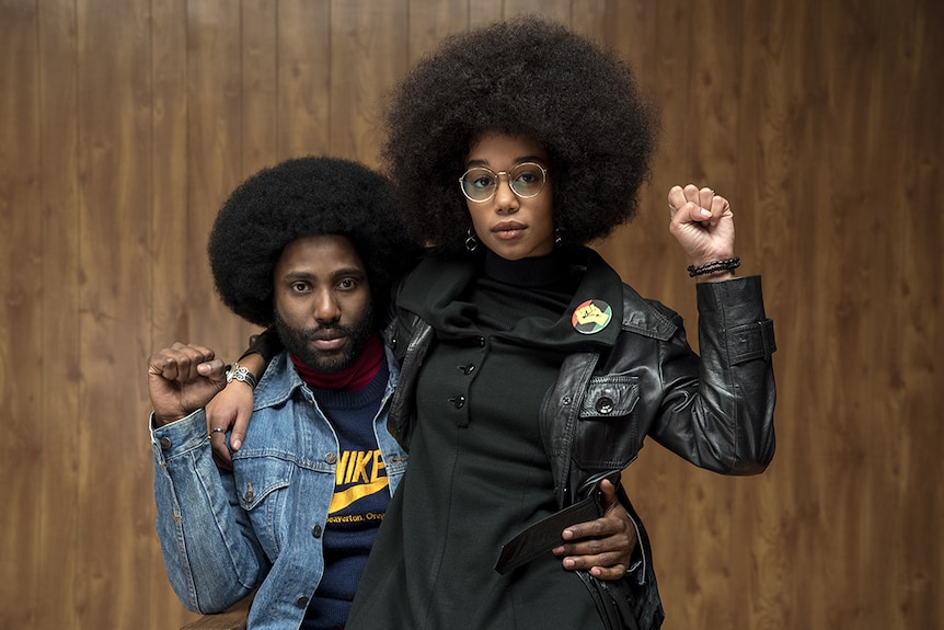 Image from 2018 film BlacKkKlansman of Laura Harrier sitting on John David Washington's knee, both with raised closed fists.