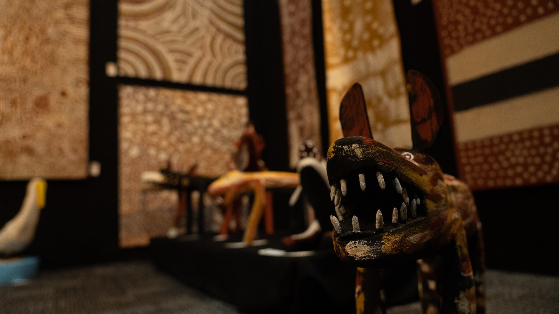 A series of sculptures in the shape of dingoes lined up on the floor of an art gallery, in front of paintings displayed on walls.