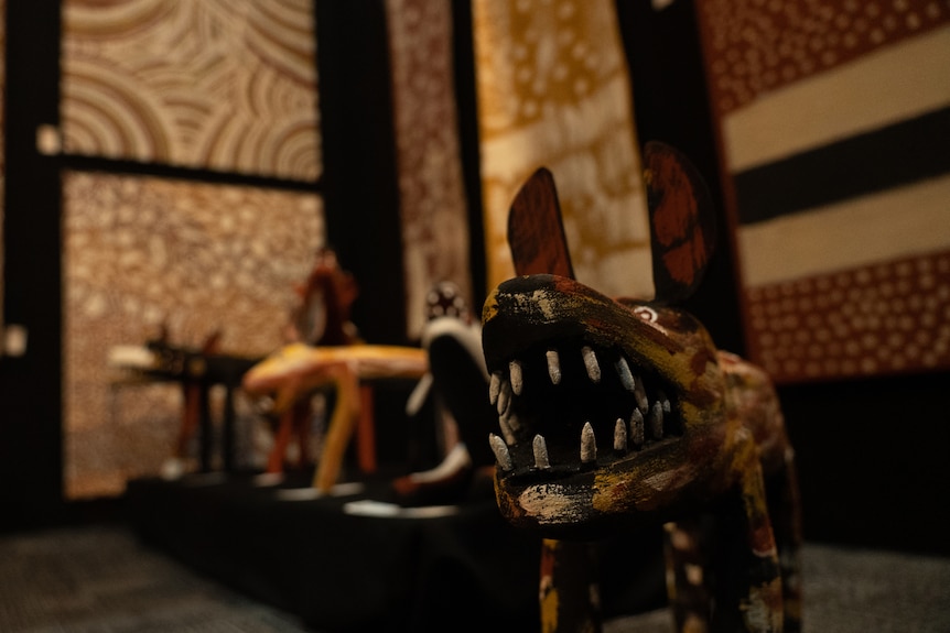 A series of sculptures in the shape of dingos lined up on the floor of an art gallery, in front of paintings displayed on walls.