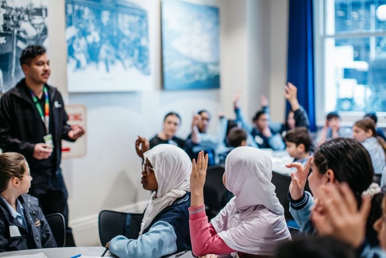 In what looks like a classroom, young students in school uniforms raise their hand and face a man at the front of the room.