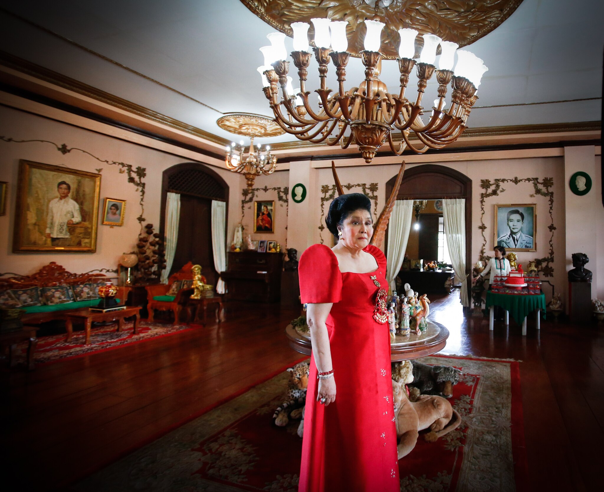 Imelda Marcos in a bright red dress adorned with jewels stands under a chandelier 