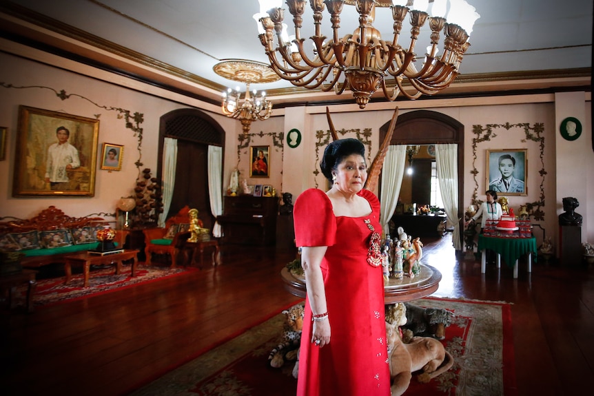Imelda Marcos in a bright red dress adorned with jewels stands under a chandelier 