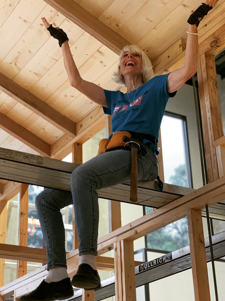 An older woman sits on an elevated wood beam in a house structure, smiling.