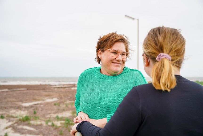 A woman wearing a green jumper and glasses, standing on a jetty, smiles at a younger woman.
