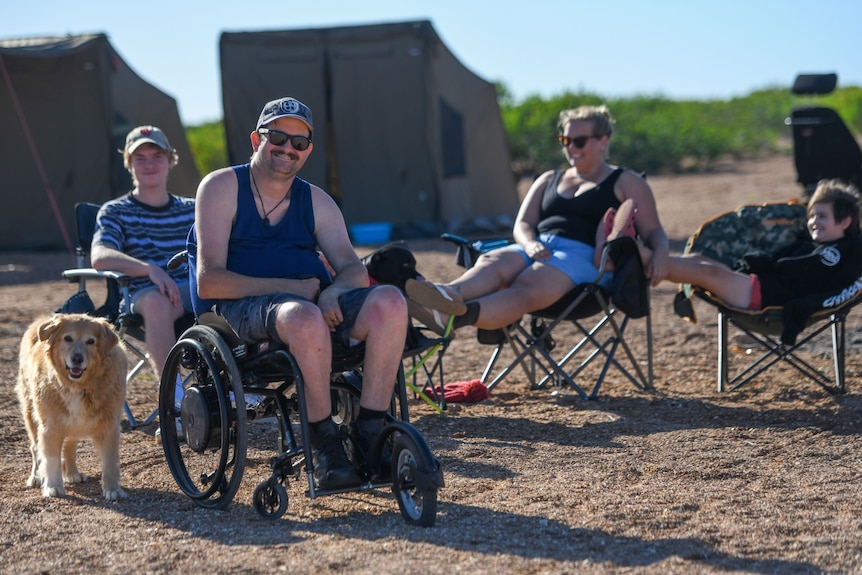 Ben Alridge sits at the camp site in his wheelchair smiling, while surrounded by his family