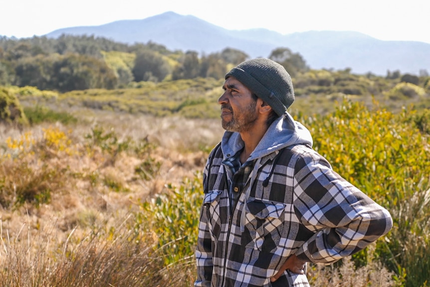 Warren Foster stands in rich open grassland with silhouette of Mount Gulaga behind him