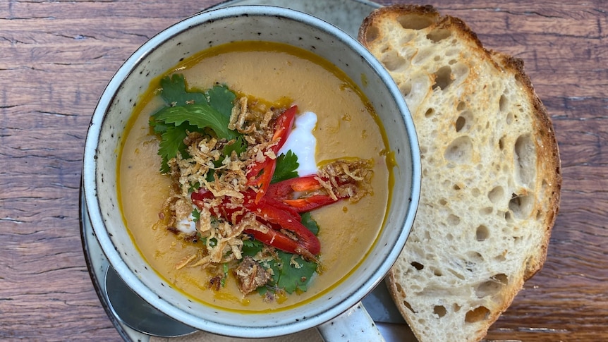 A mug of orange-coloured soup with chilli and coriander garnish, on a plate with bread on a wooden table