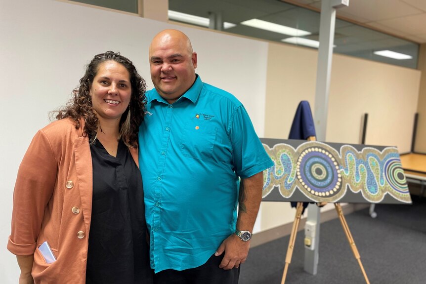 A woman and a man stand in front of an Indigenous artwork.