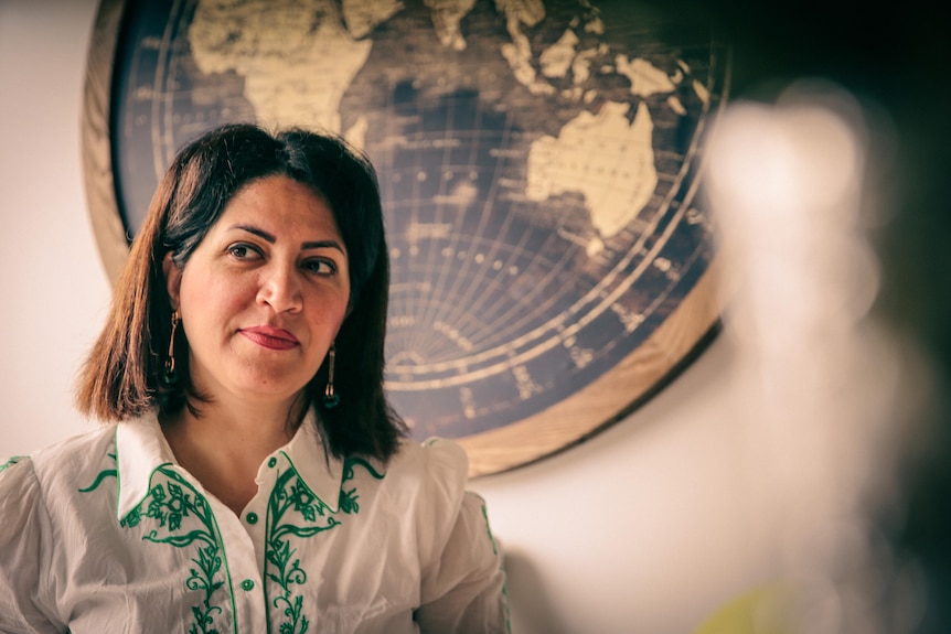 A woman with shoulder-length hair stands in front of an atlas, looking to her right.