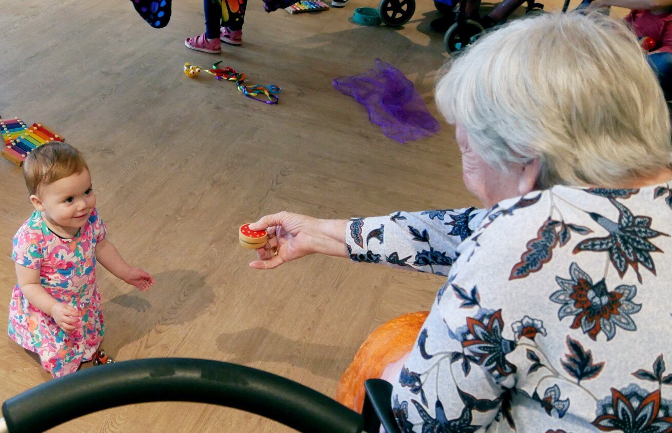 A baby looks up at an elderly woman who is holding out a small music shaker