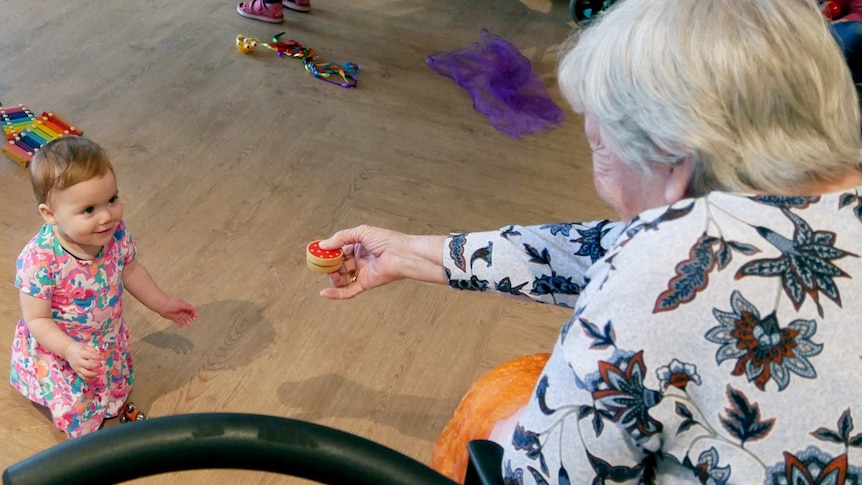 A baby looks up at an elderly woman who is holding out a small music shaker