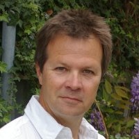 A man wearing a white shirt in front of a garden.