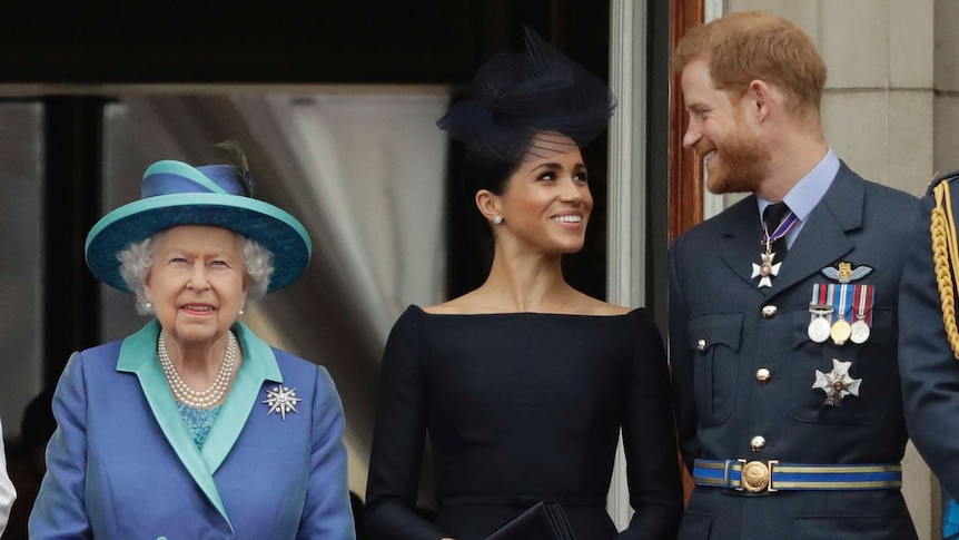 Queen Elizabeth II stands on a balcony, while Harry and Meghan, Duke and Duchess of Sussex, look at each other.