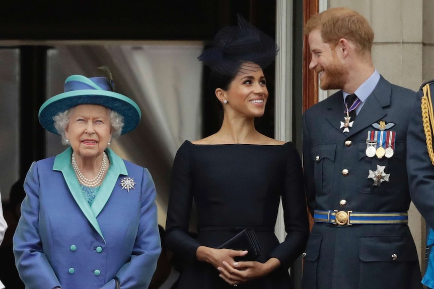 Queen Elizabeth II stands on a balcony, while Harry and Meghan, Duke and Duchess of Sussex, look at each other.