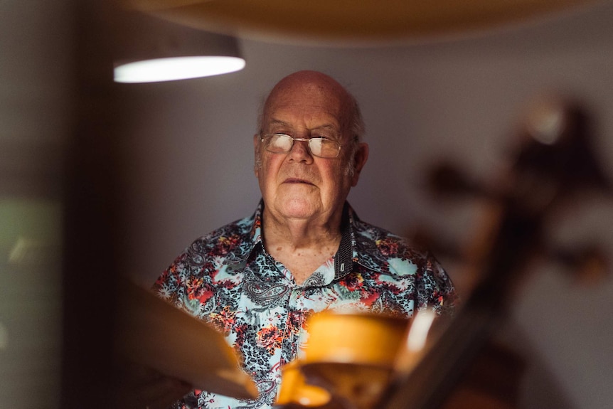 A bald man wearing a dark floral shirt sits down at a desk tuning up a violin