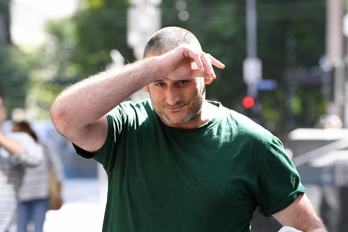 A man with a buzz cut and wearing a green t-shirt walks outside court on a sunny day with his hand on his forehead.