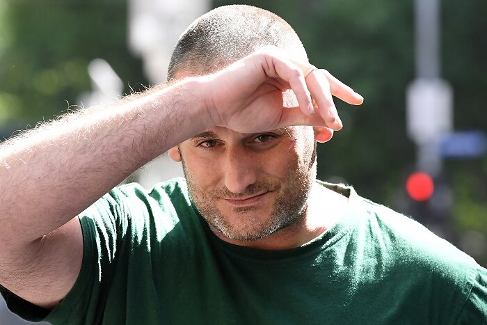 A man with a buzz cut and wearing a green t-shirt walks outside court on a sunny day with his hand on his forehead.