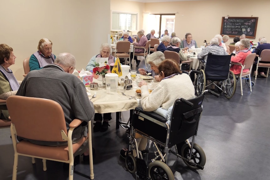 A group of elderly people sit at tables in a care home eating lunch