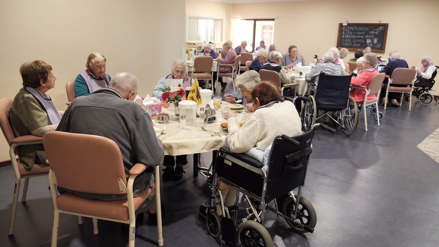 A group of elderly people sit at tables in a care home eating lunch