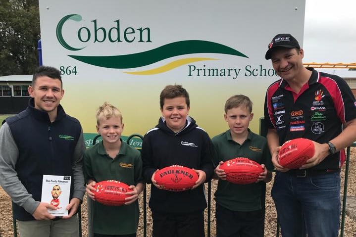 Two men and three primary school students hold footballs in front of the Cobden Primary School sign.