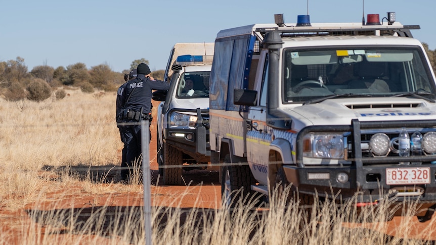 Two four-wheel-drive police cars are seen behind a wire fence with two police officers standing at one car,