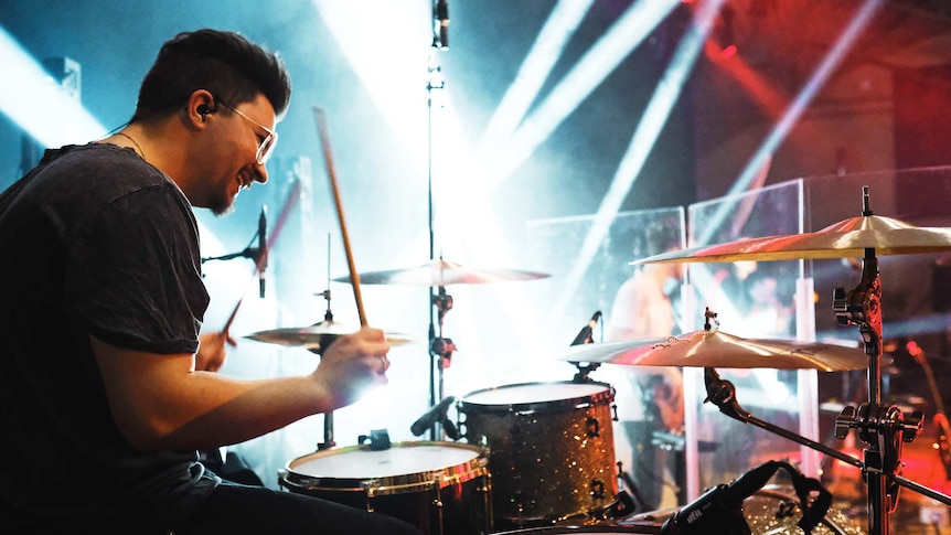 Smiling man playing drums on stage with blue and red lights.