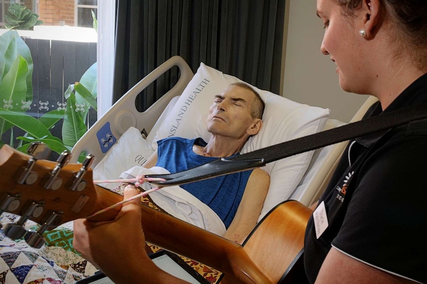 A man in a hospital bed listens to a woman playing a guitar