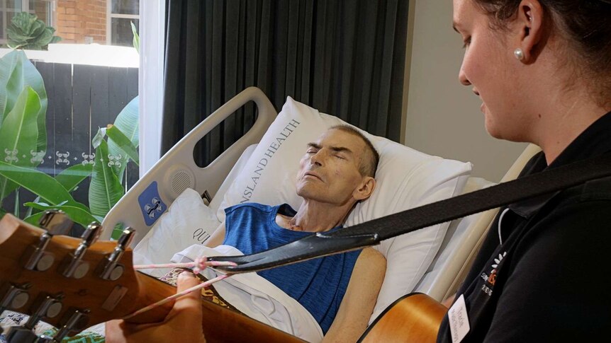 A man in a hospital bed listens to a woman playing a guitar