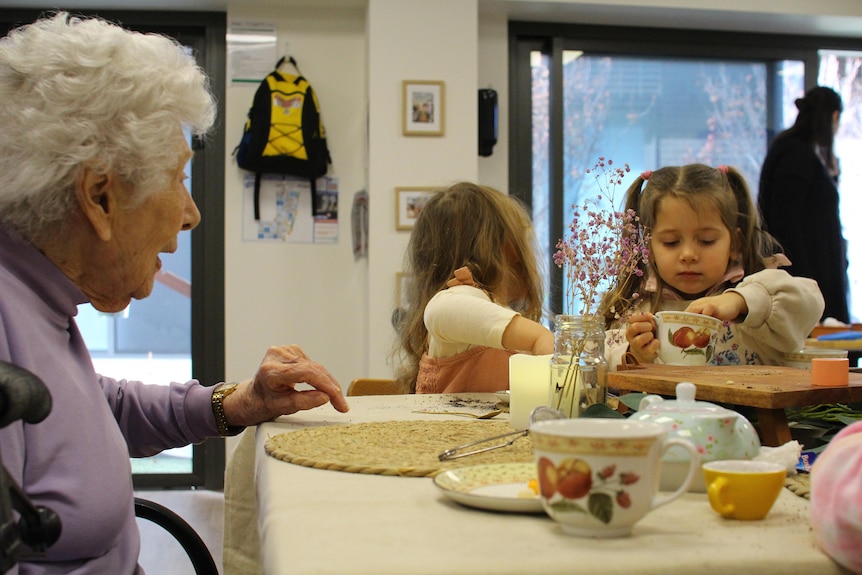 AN elderly woman sits at a table with two small girls making tea