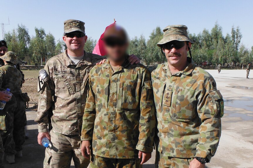 Three men in army uniform smile in a photo outdoors. The man in the centre's face is blurred.