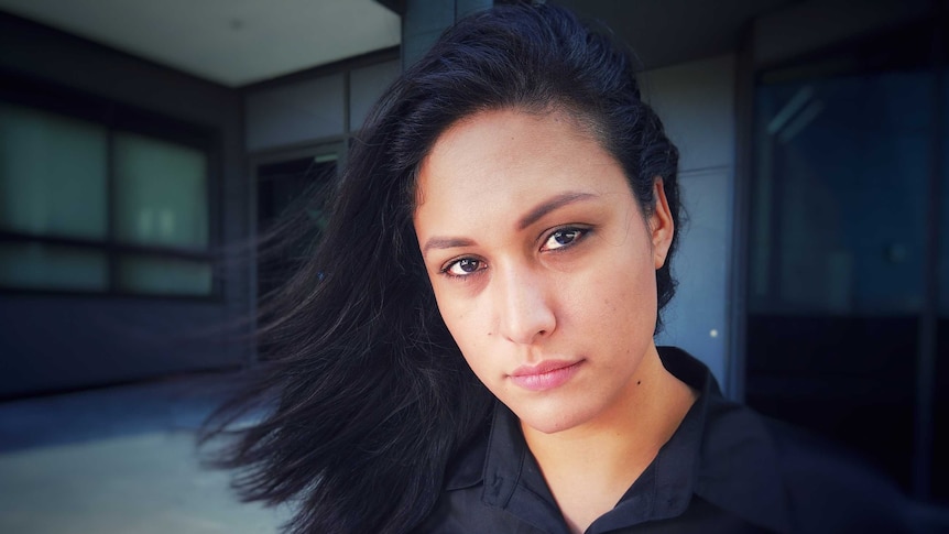 A woman with dark wind-blown hair looks directly and seriously into the camera.