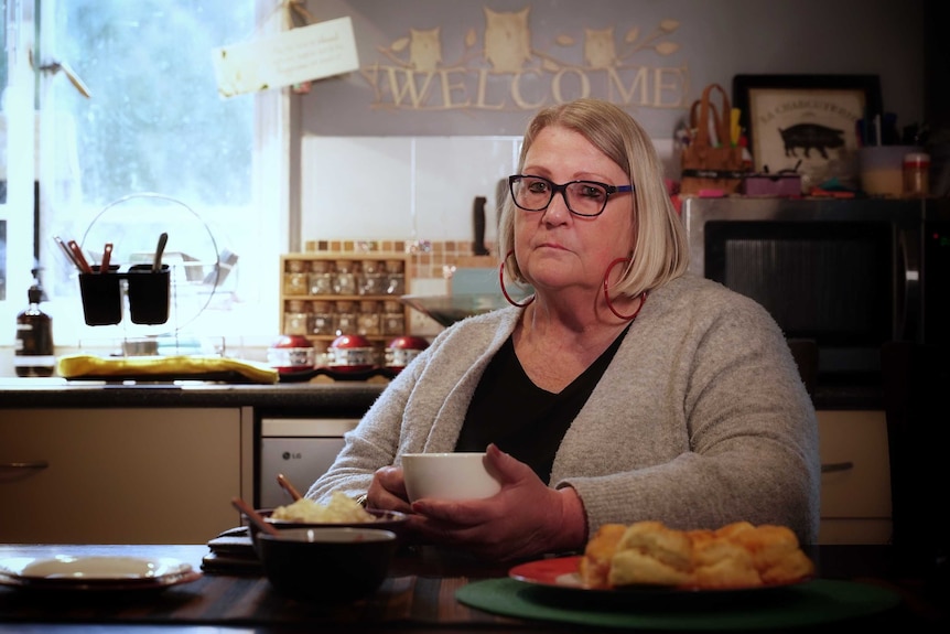 Kathy Flynn sits in her kitchen at a table of scones and holds a cup of tea.