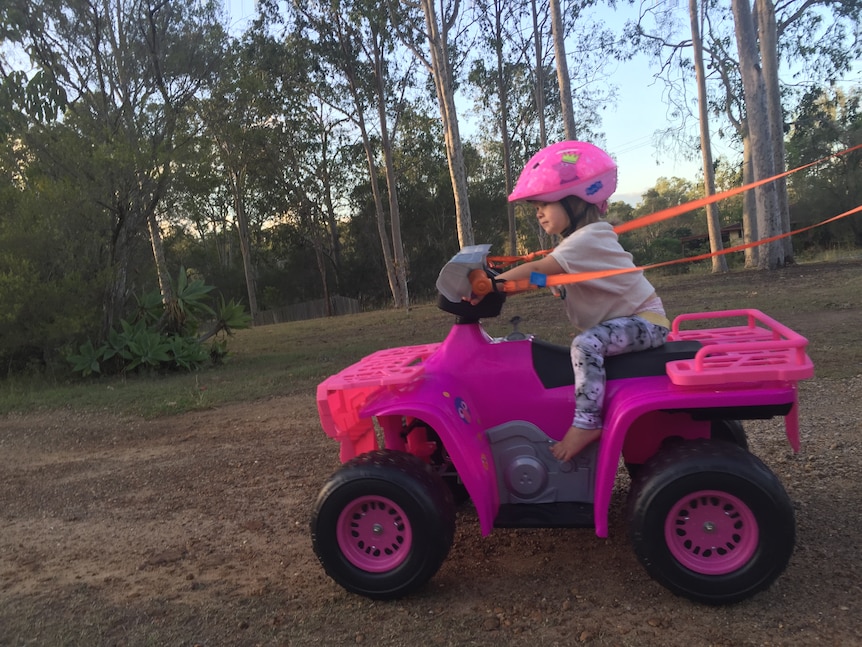 A one-year-old girl drives a pink quad bike with straps attached for steering.