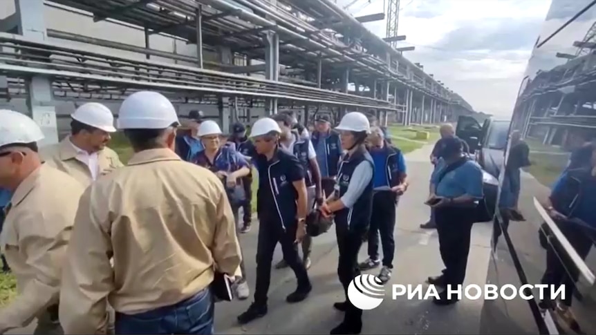 IAEA officials in navy vets wear white helmets as they are shown around a nuclear power plant.