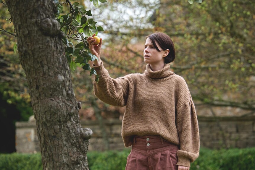 White woman with short brown hair wearing taupe jumper and rouge pants stands in garden holds and looks at a lemon on a tree