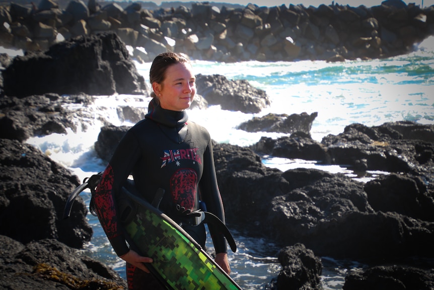 A young woman in a black wetsuit, carrying a green surfboard. She is standing on coffee rocks as waves break nearby