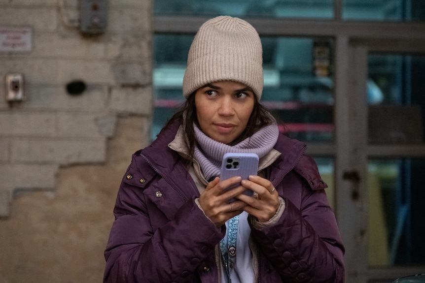 A Latina woman in the 40s wears a big purple snow jacket and a cream beanie and looks off camera while holding a phone.