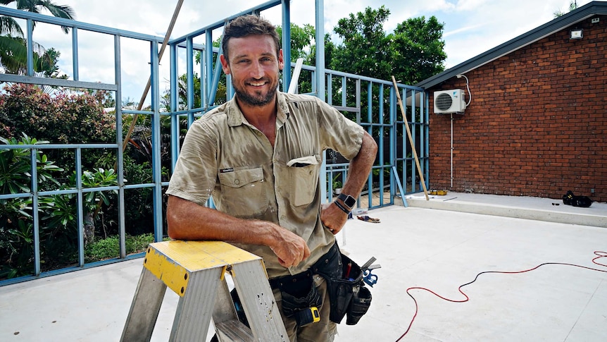 Peter MacFarlane is at a building site smiling at the camera. He is leaning on a step ladder and wearing a tool belt.