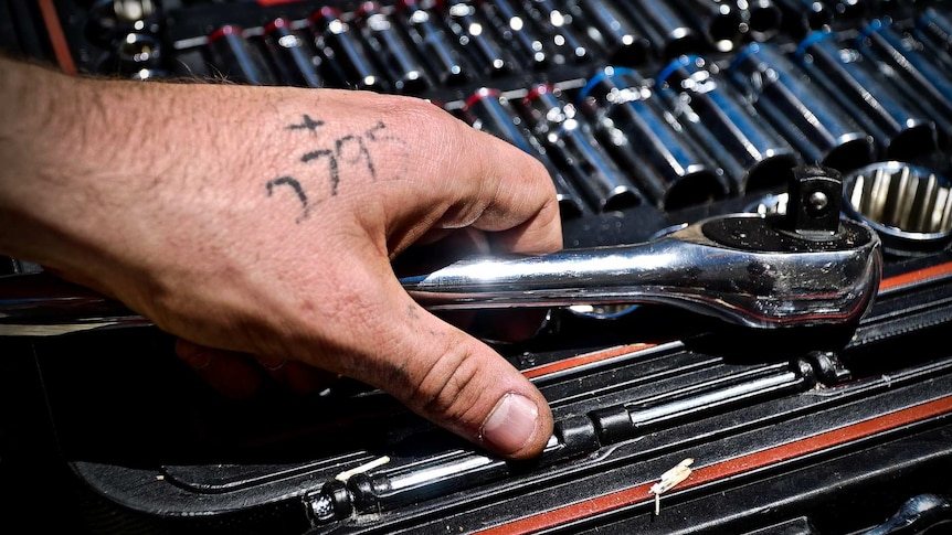 A hand holding a wrench shows a four-digit tattoo.