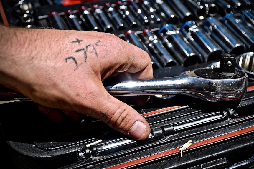 A hand holding a wrench shows a four-digit tattoo.
