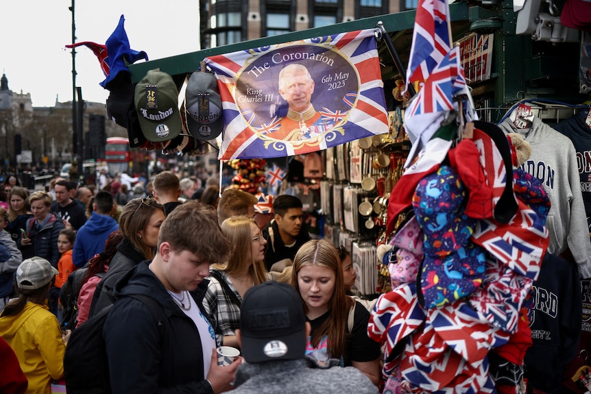 Crowds of people stand near a street kiosk selling flags, hats, jumpers and other merchandise featuring King Charles