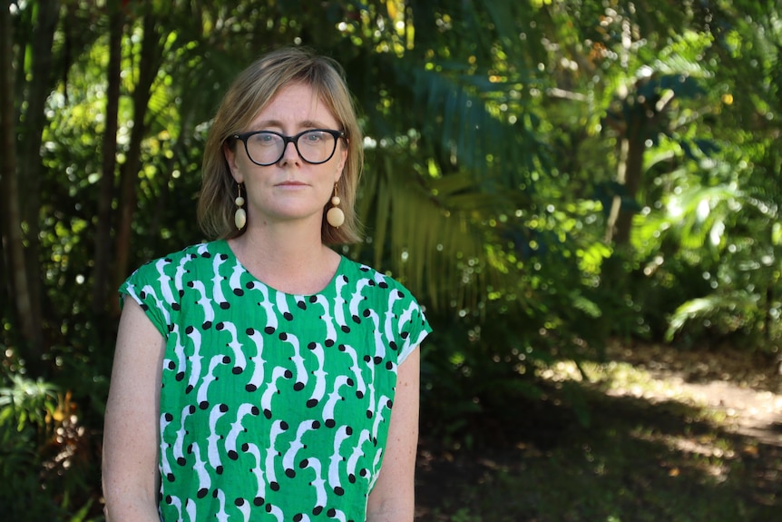 A woman in glasses and a green shirt looks into the camera. She is in a leafy outdoor space. 