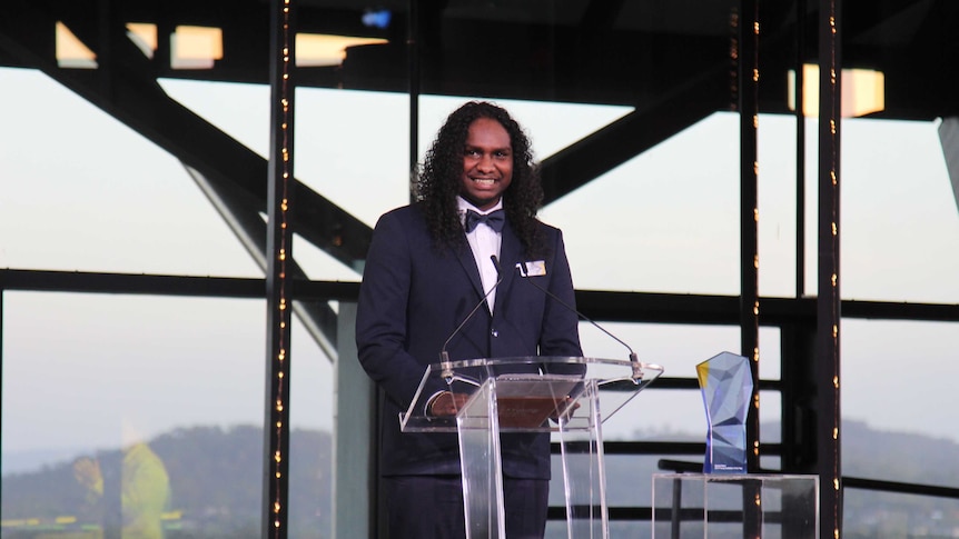 Baker Boy, in a dark suit, stands at a podium to accept his Young Australian of the Year award for 2019.