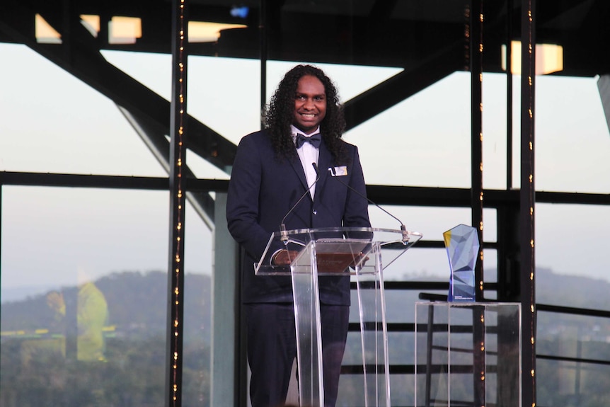 Baker Boy, in a dark suit, stands at a podium to accept his Young Australian of the Year award for 2019.